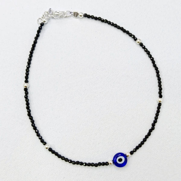 Anklet with black beads and a blue evil eye charm on a light gray background