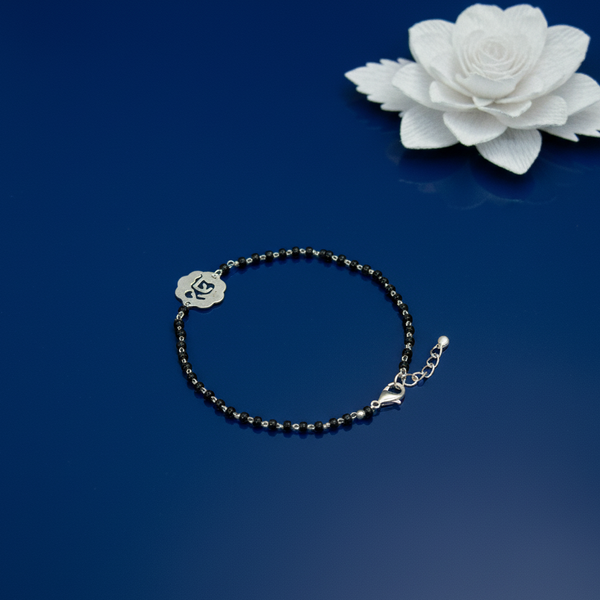 Black beaded bracelet with a silver flower charm placed on a blue reflective surface beside a white decorative flower.